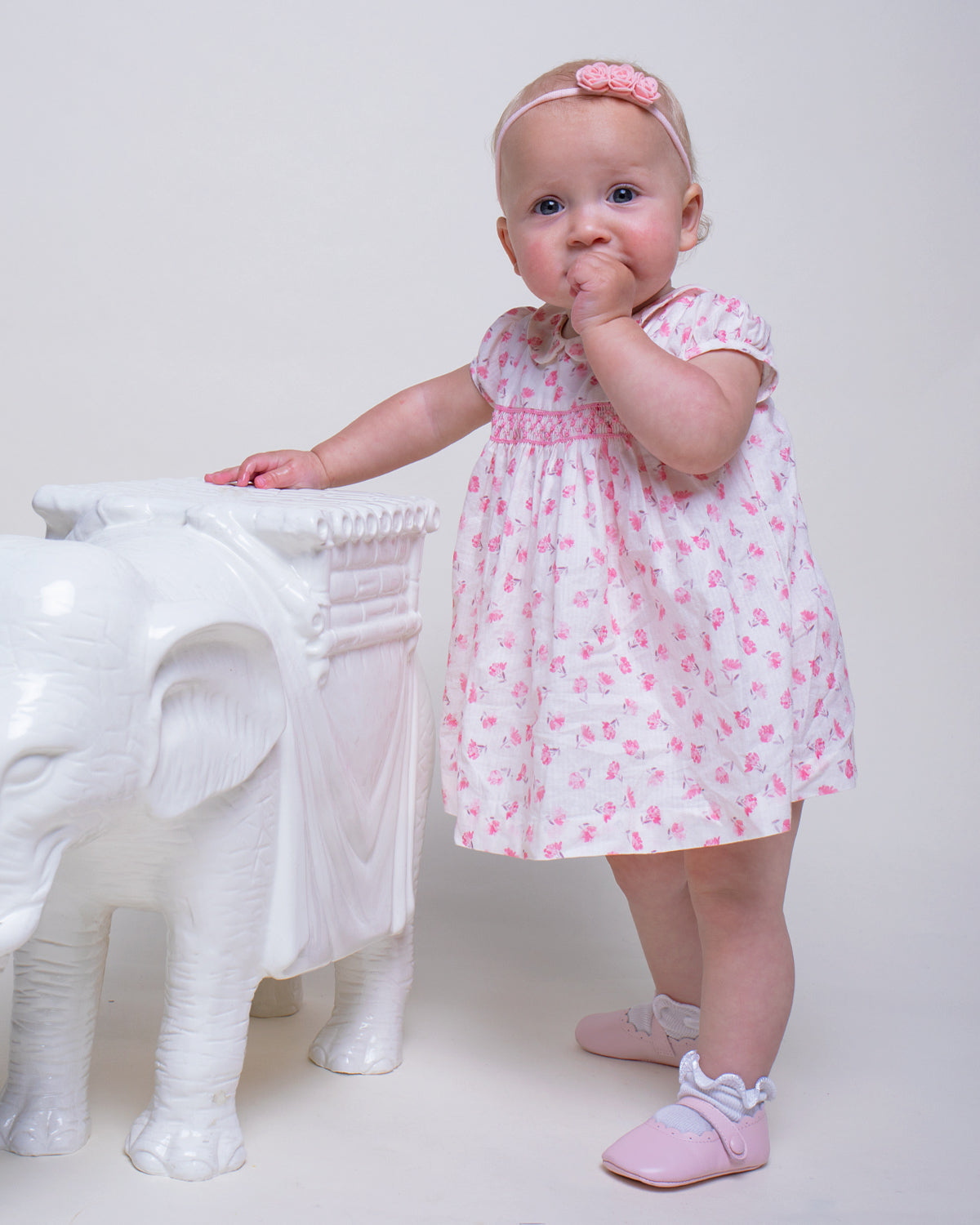 Toddler model wearing the Baby Eva Versaille Rose Octuouse dress set while standing next to a white elephant pedestal, styled with matching headband and shoes.