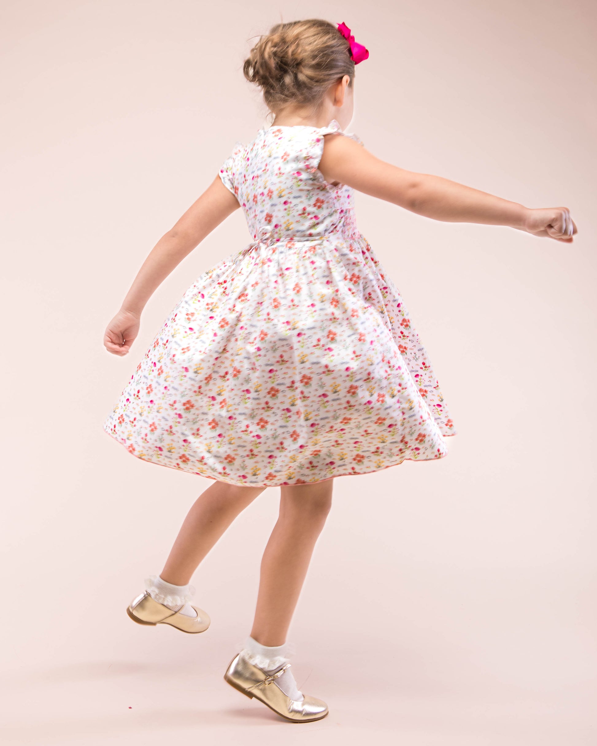 Girl wearing Bonnie Happy Garden Smocked Dress with gold shoes and pink hair bow moving- showing the back and the side of the dress