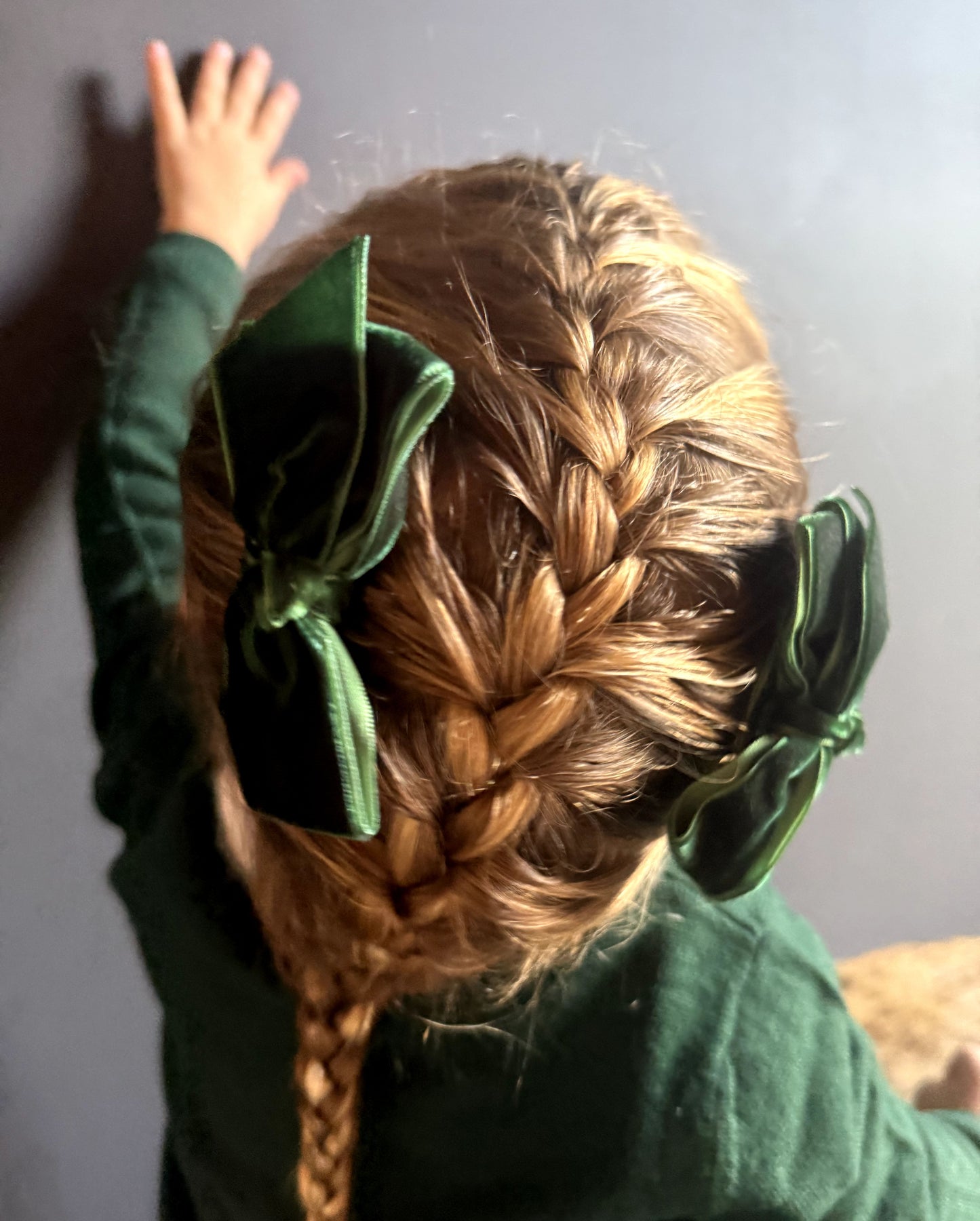 Little girl with braided hair wearing green velvet hair bows, green sweater facing away from camera  against a plain background