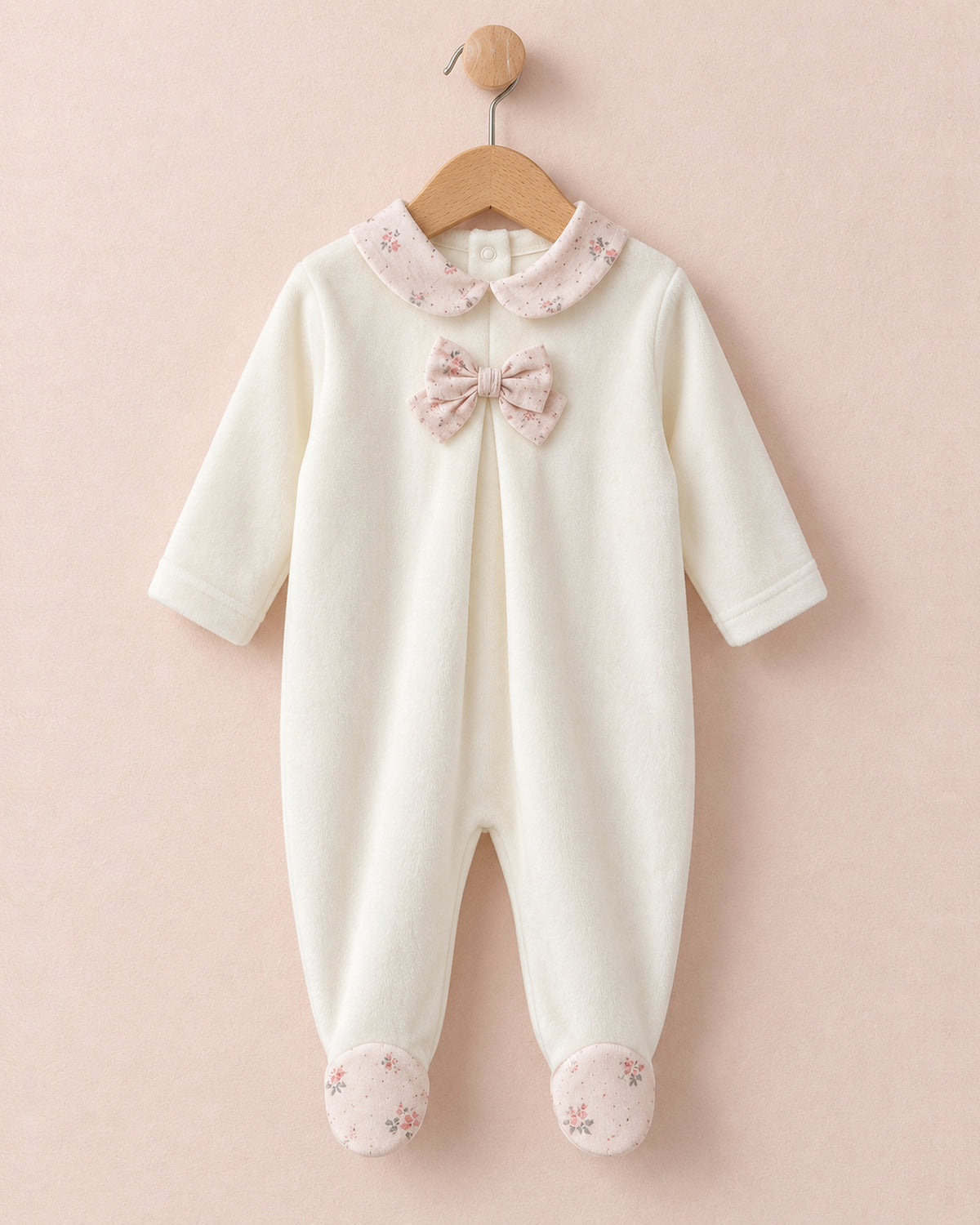 Front of white velour baby onesie hanging from wooden hanger, with light pink bows in center of chest, and pink floral motif collars and feet.