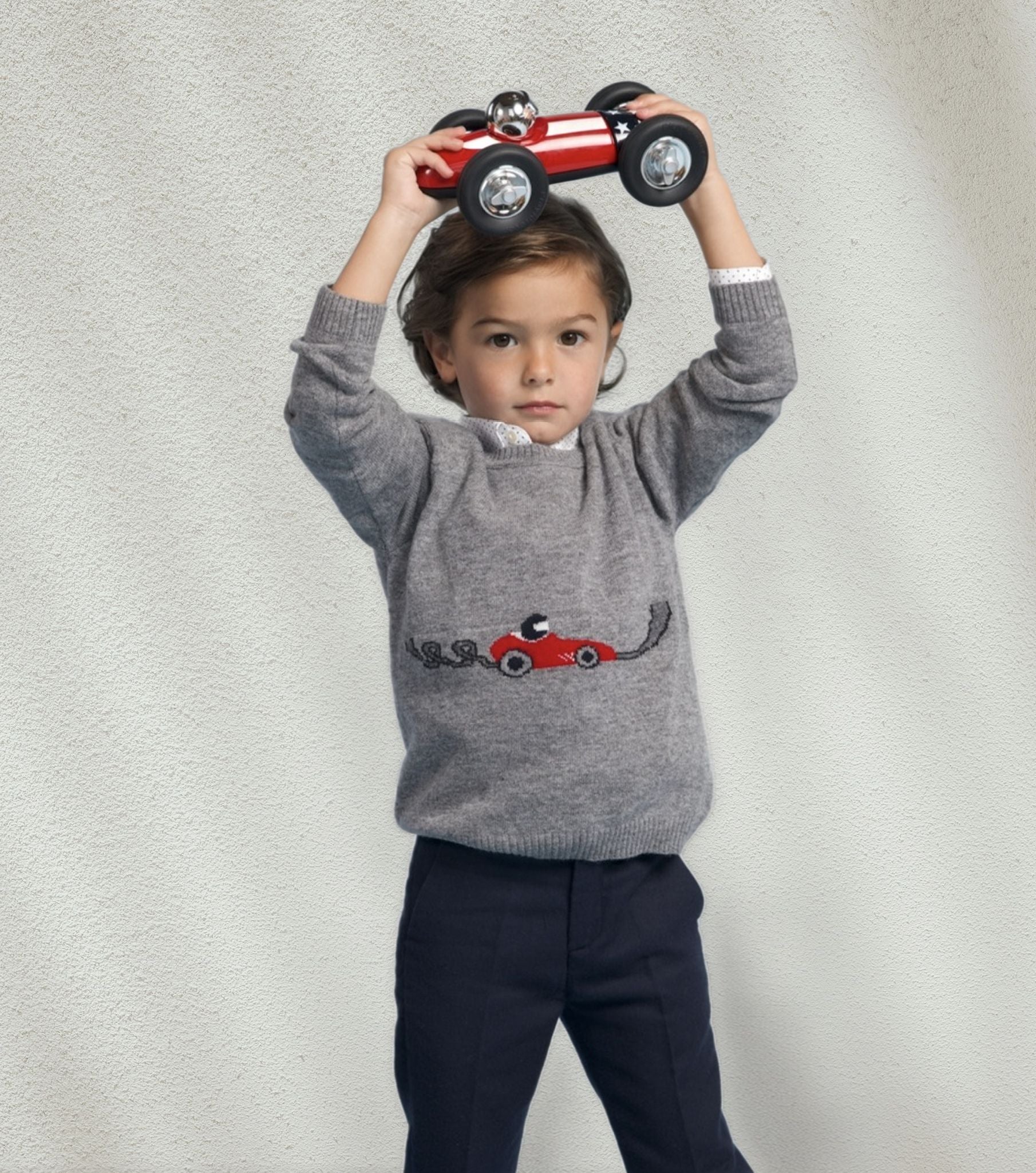 Child wearing a gray sweater with a red car design, holding a small red car above their head against a light gray background.