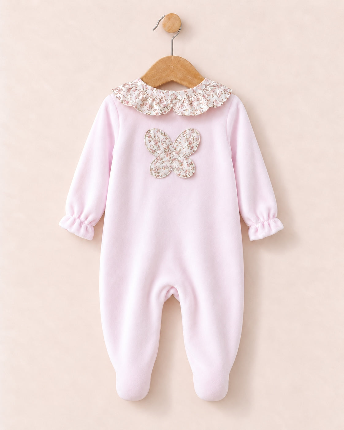 A baby footed one-piece jumpsuit in light pink, featuring a decorative floral lace ruffled collar and a matching floral applique butterfly on the chest, hanging on a wooden hanger against a solid pale pink background.