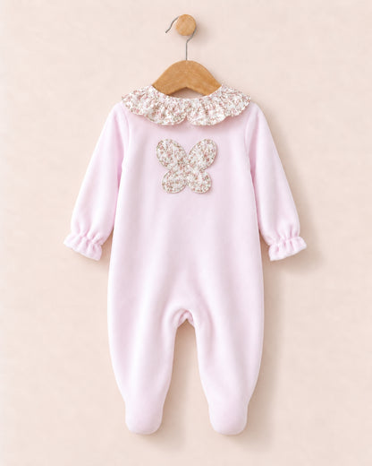 A baby footed one-piece jumpsuit in light pink, featuring a decorative floral lace ruffled collar and a matching floral applique butterfly on the chest, hanging on a wooden hanger against a solid pale pink background.