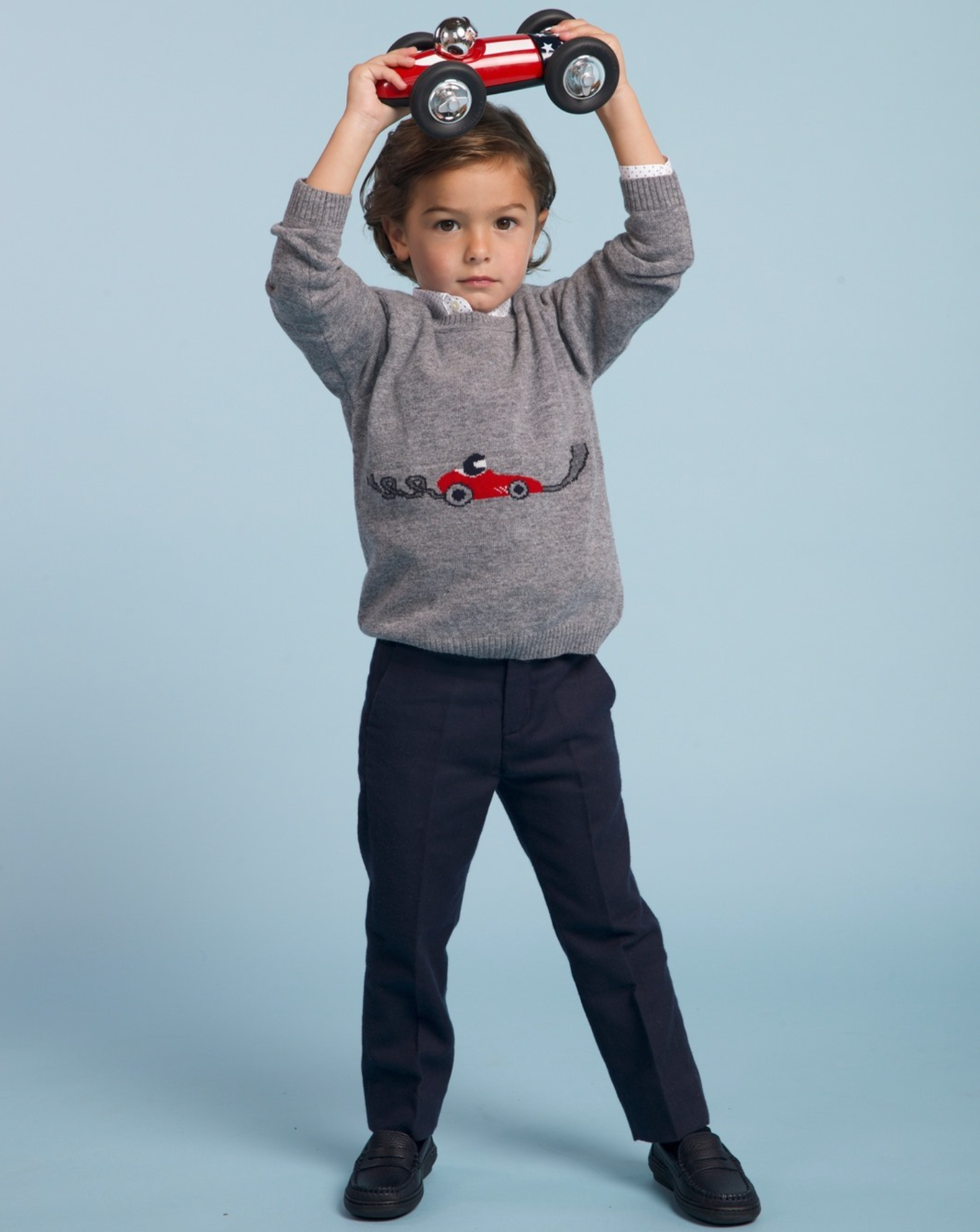 Child wearing a gray sweater with a red car design, holding a small red car above their head against a light blue background.