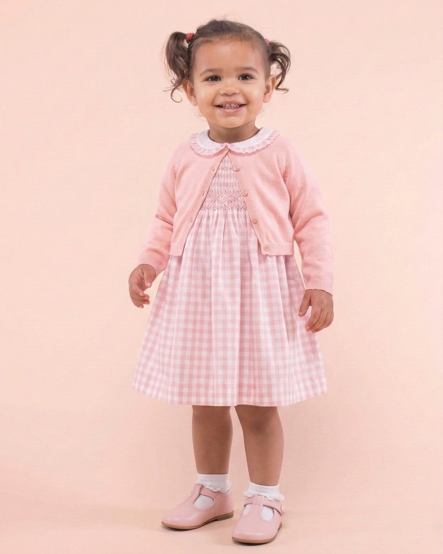 Young girl wearing a pink checkered dress with a cardigan against a light pink background