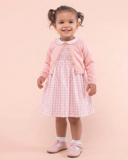 Young girl wearing a pink checkered dress with a cardigan against a light pink background