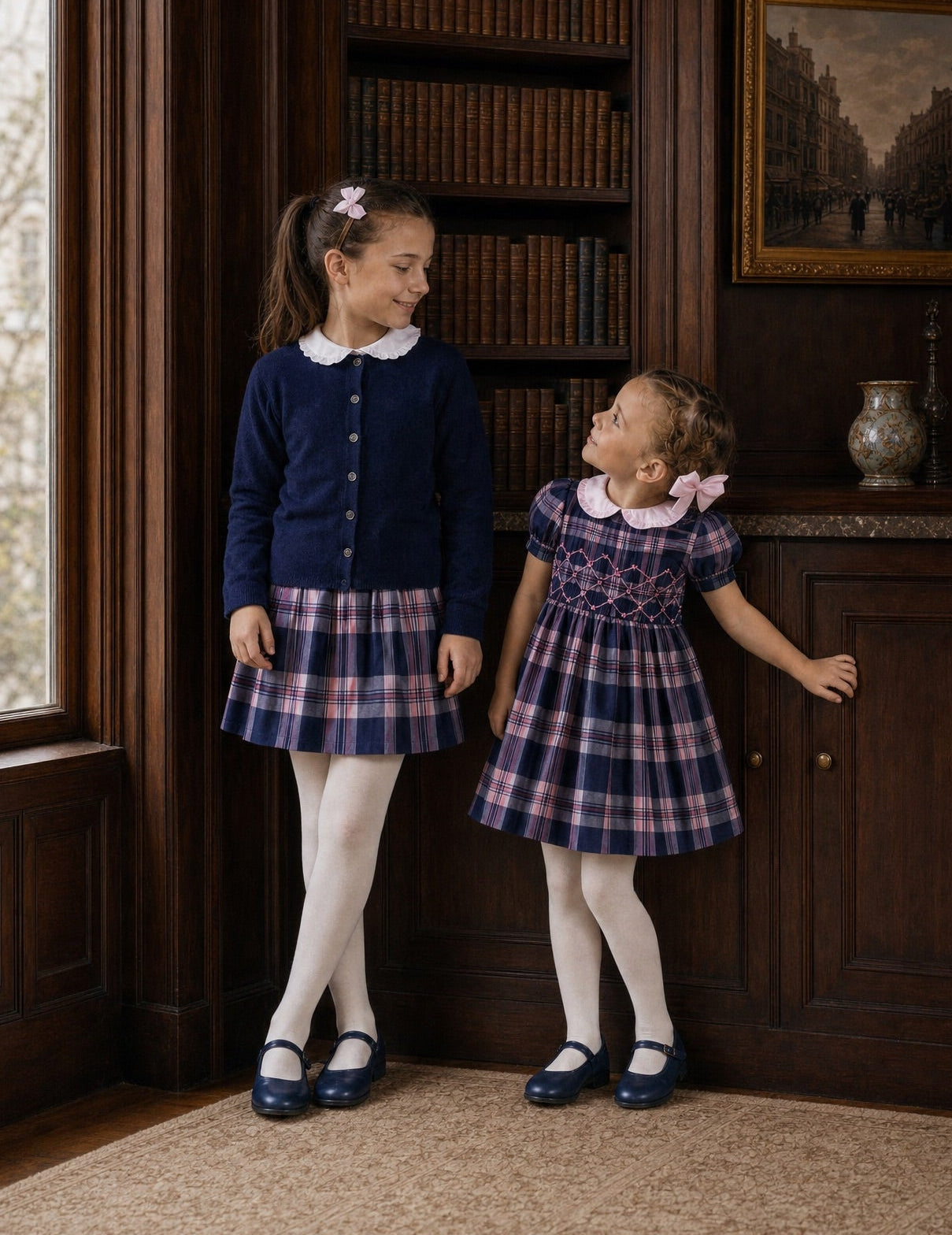 Two young girls standing in a classic library wearing matching navy blue and red plaid school uniform dresses with white Peter Pan collars and white tights.