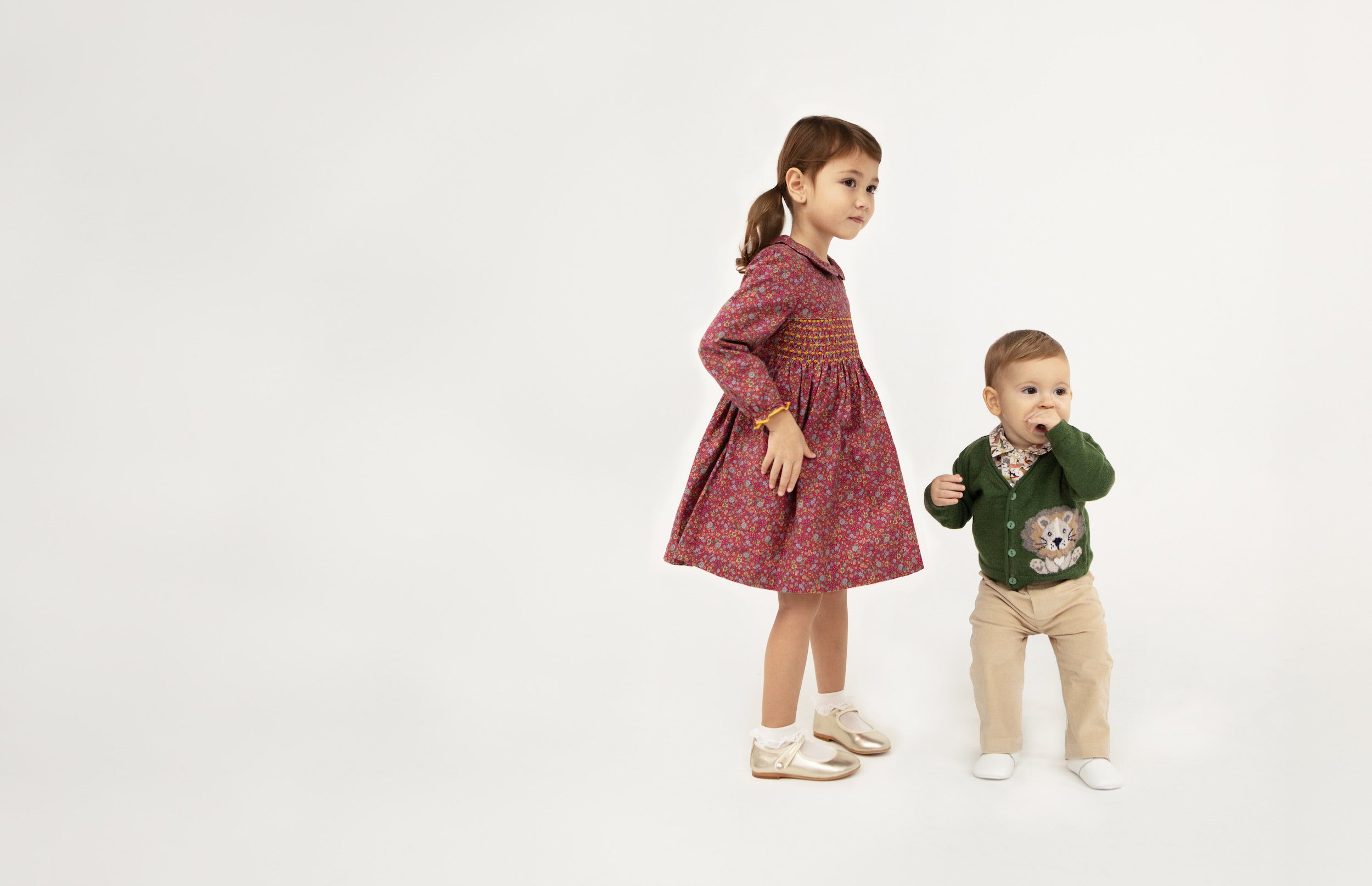 One girl wearing a dark red dress and a boy wearing a green sweater with a lion, and tan pants, against a white background