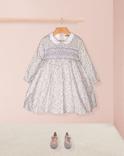 A styled front-facing shot of the Fiorella Belina dress alone, laid flat with coordinating navy blue Mary Jane shoes, emphasizing its full, twirl-ready skirt and smocking details.