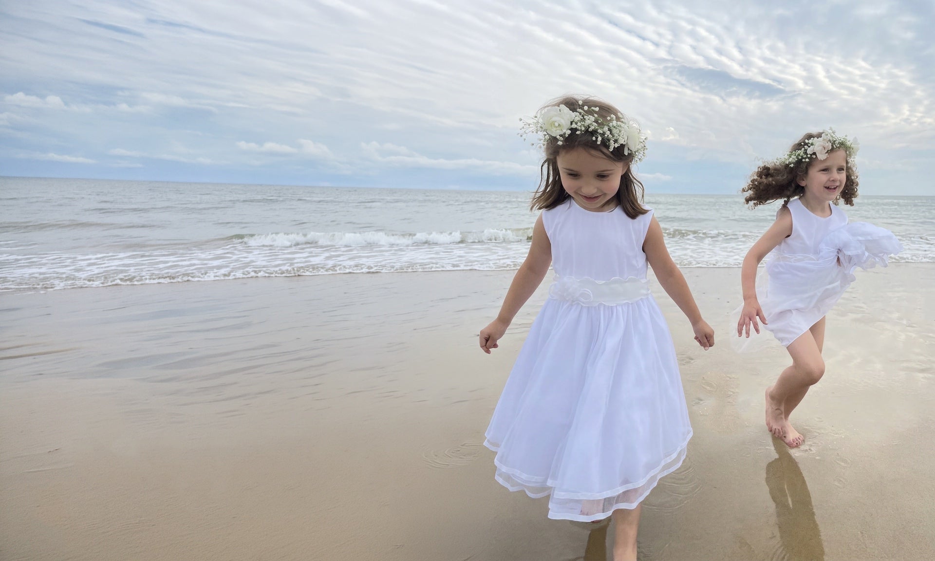 Two young girls in white dresses running on a beach with ocean waves in the background.