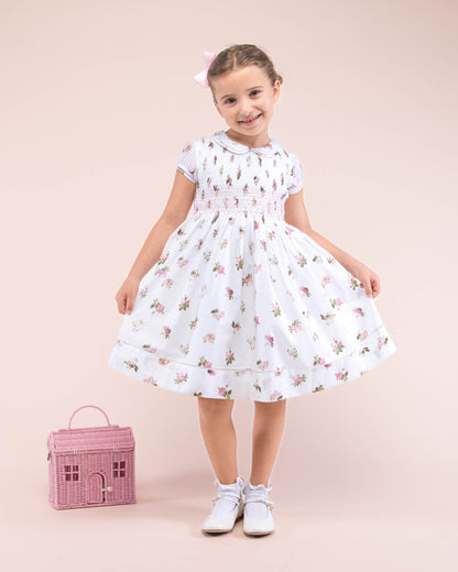 Girl model standing, wearing Emma Delicate Blush Rose Smock Dress showing the front of the dress with a Pink bag on the floor to match the outfit 