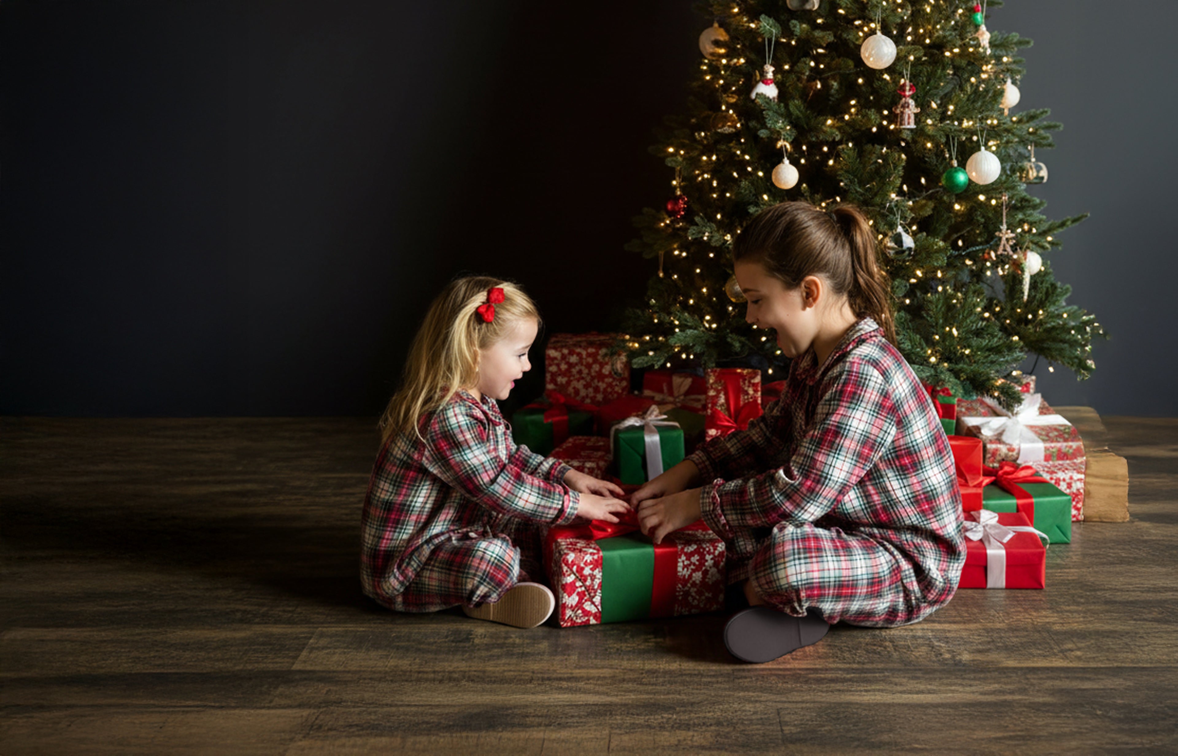 Two children in plaid pajamas opening Christmas presents in front of a decorated tree.