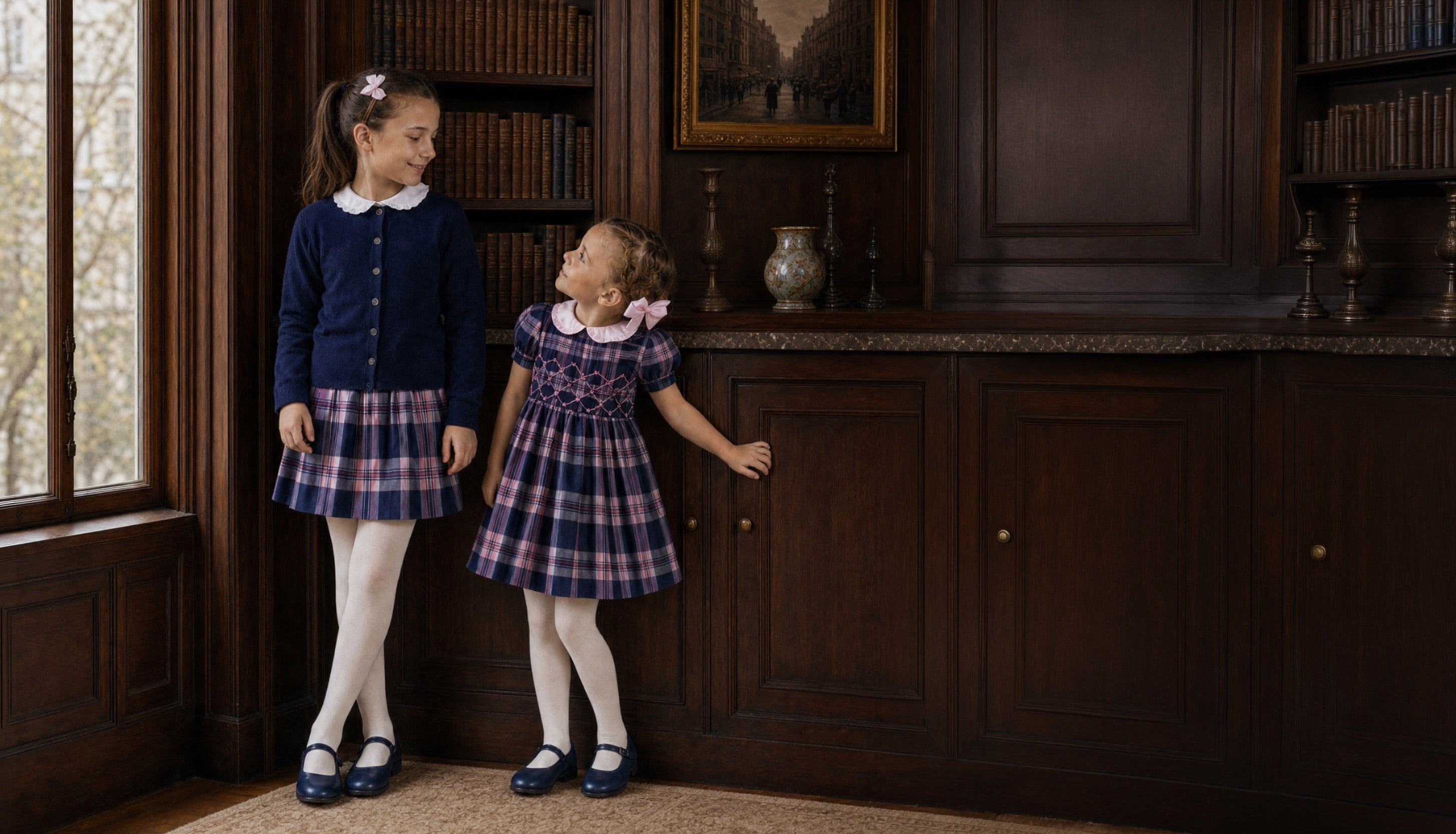 Two young girls standing in a dark wood library or study, wearing coordinated navy and plaid outfits. One girl wears a navy cardigan with a white collar and plaid skirt, and the other wears a matching navy and plaid dress with a white collar.