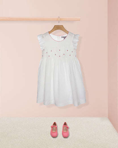 White dress with red embellishments on a hanger against a pink background, with pink shoes on the floor.