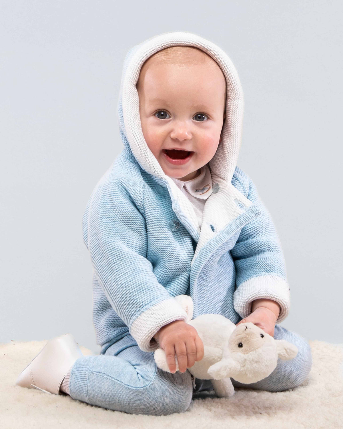 A baby sitting on a cream carpet wearing a light blue hooded knit romper with white trim, holding a small white plush toy against a light gray studio background.