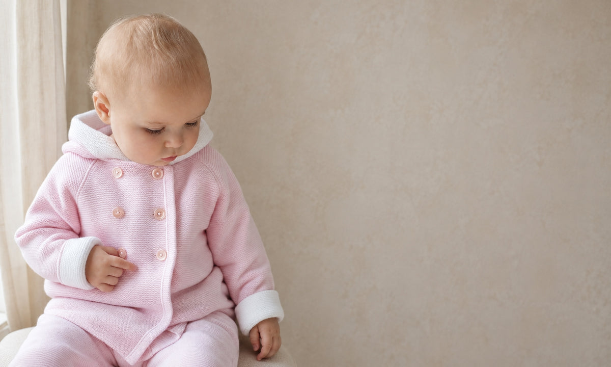 A baby wearing a light pink hooded cardigan and matching pants with white cuffs, sitting against a neutral beige wall.