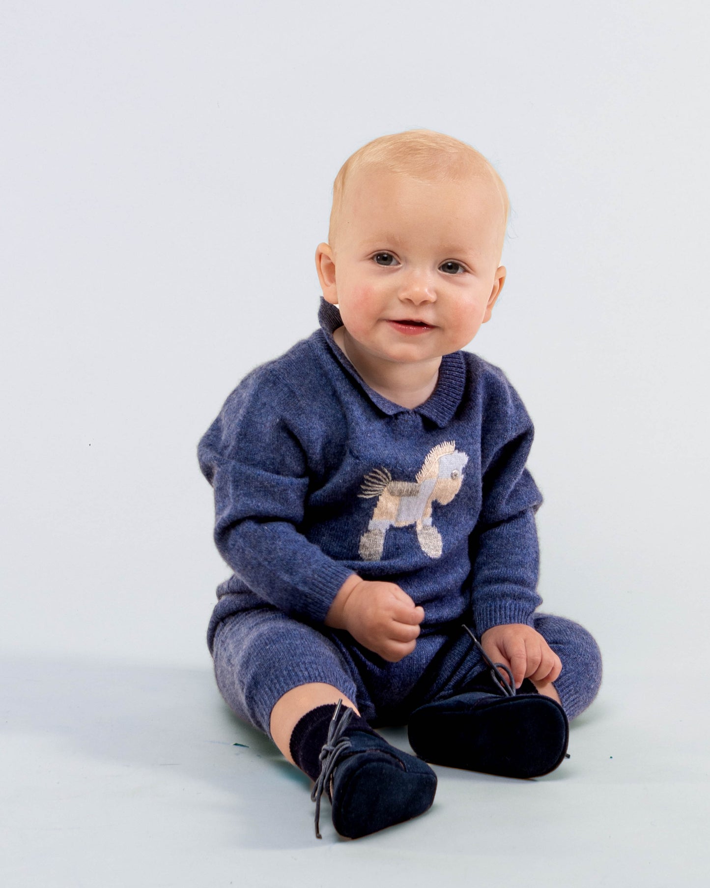 Baby boy model sitting down, smiling- wearing the Logan Jean Cashmere Romper showing the front of the romper
