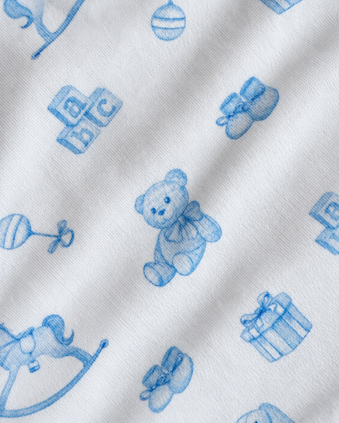 White fabric with a blue repeating pattern of baby-themed illustrations including a teddy bear, blocks, booties, a rattle, a rocking horse, and a gift box.