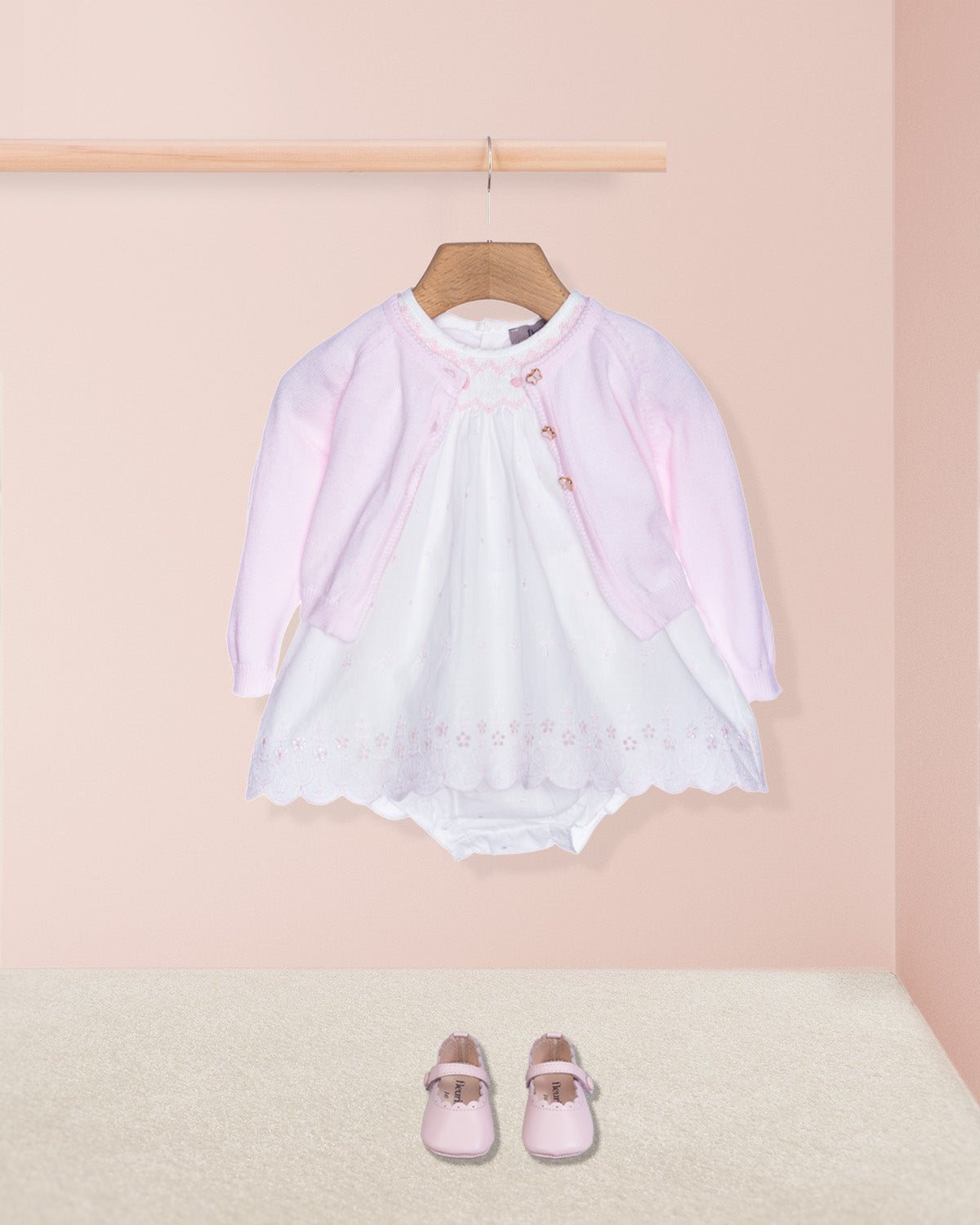 Baby outfit with pink cardigan, white dress, and shoes on a pink background