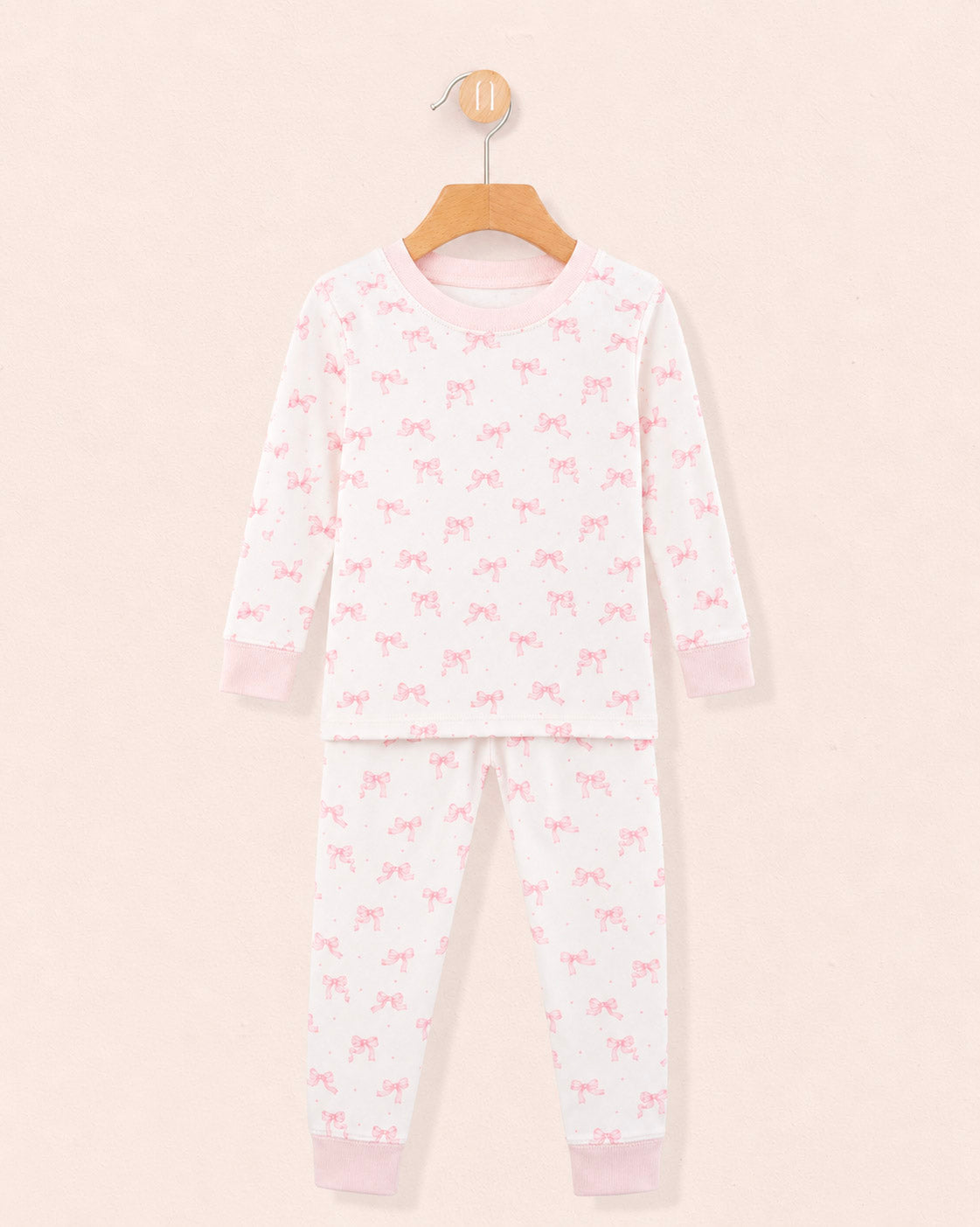 white pajamas with pink bows all over the top and bottom, pink crewneck band and pink cuffs on the wrists and ankles, hanging on a wooden hanger against a peach neutral background.