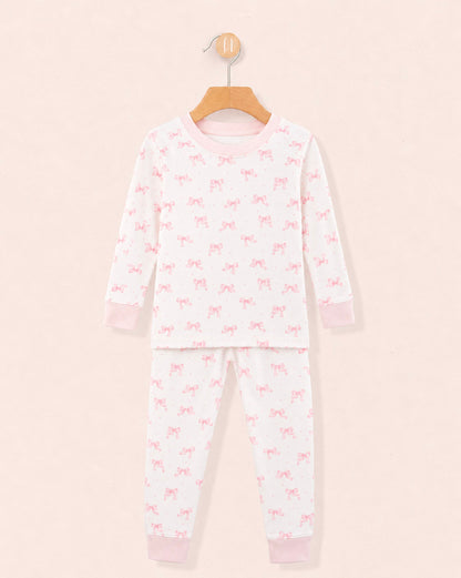 white pajamas with pink bows all over the top and bottom, pink crewneck band and pink cuffs on the wrists and ankles, hanging on a wooden hanger against a peach neutral background.