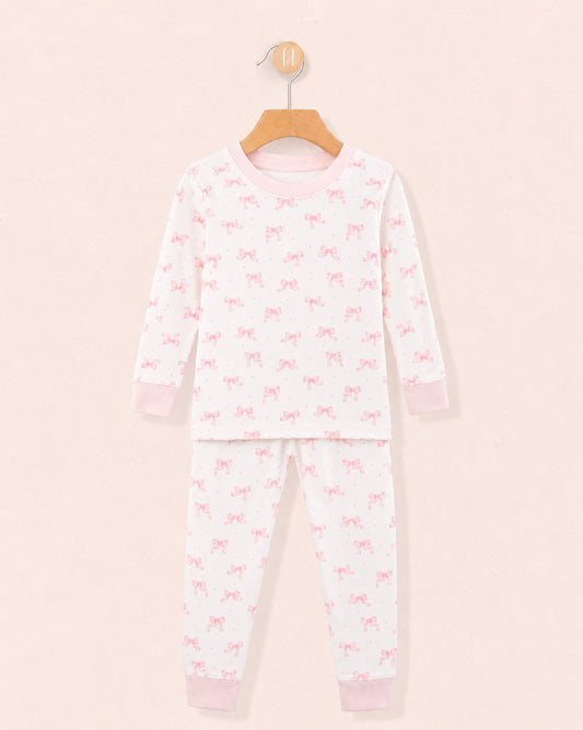 white pajamas with pink bows all over the top and bottom, pink crewneck band and pink cuffs on the wrists and ankles, hanging on a wooden hanger against a peach neutral background.