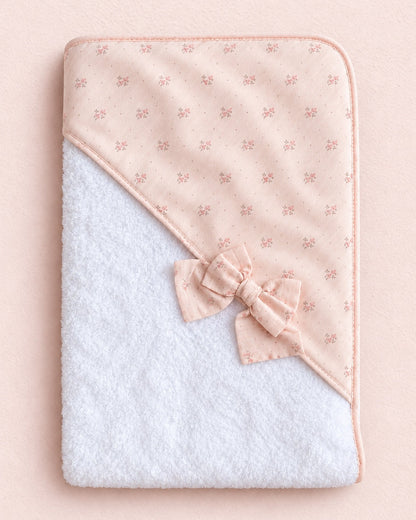 A baby hooded towel featuring a soft white terry cloth body and a light pink patterned flap with a matching bow, laid flat on a pale pink background.