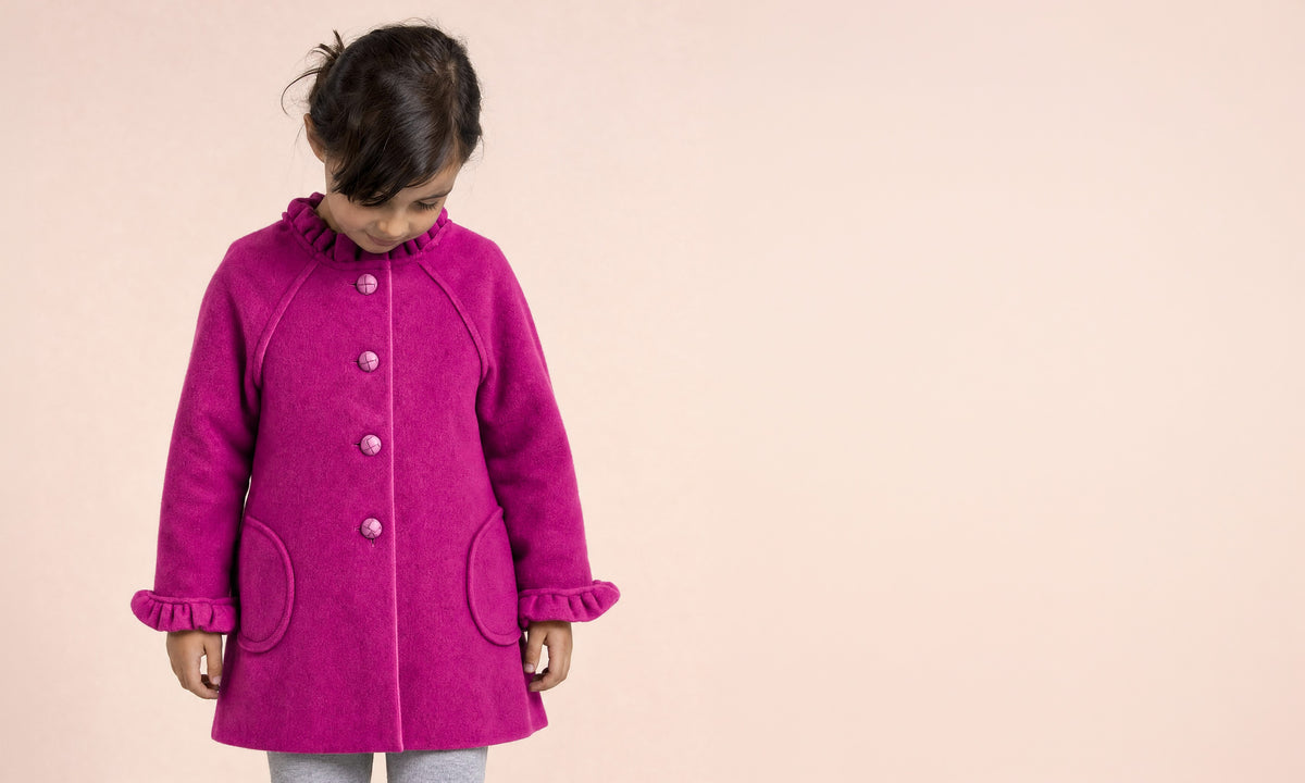 A young girl wearing a bright magenta pink wool coat with ruffled trim and front buttons, standing against a solid pale peach background.