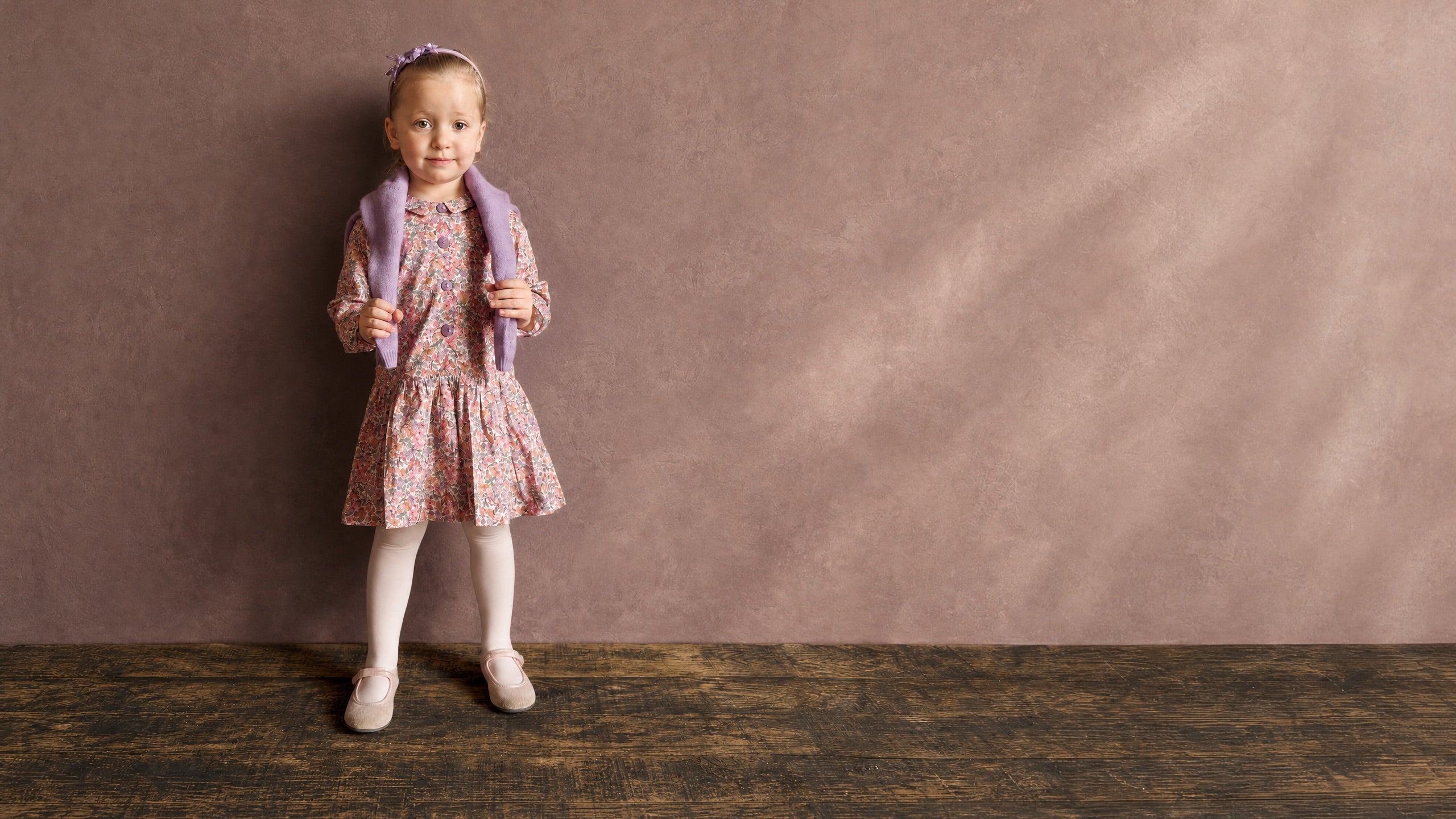 A young girl stands against a textured mauve-brown studio backdrop wearing a floral pink dress, white tights, ballet flats, and a lavender cardigan. The image has a large negative space on the right side.
