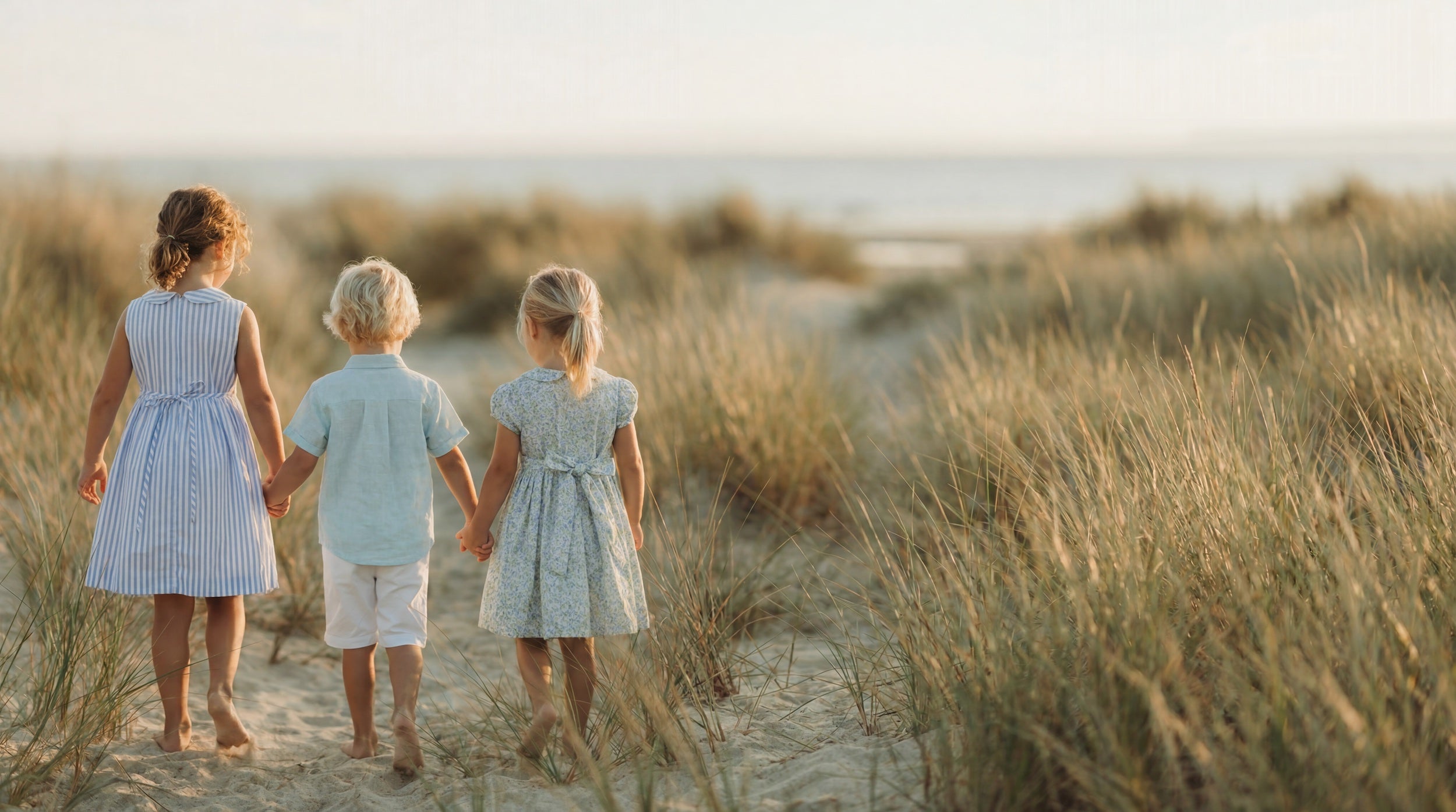 Three children holding hands and walking along a sandy path in a grassy field.