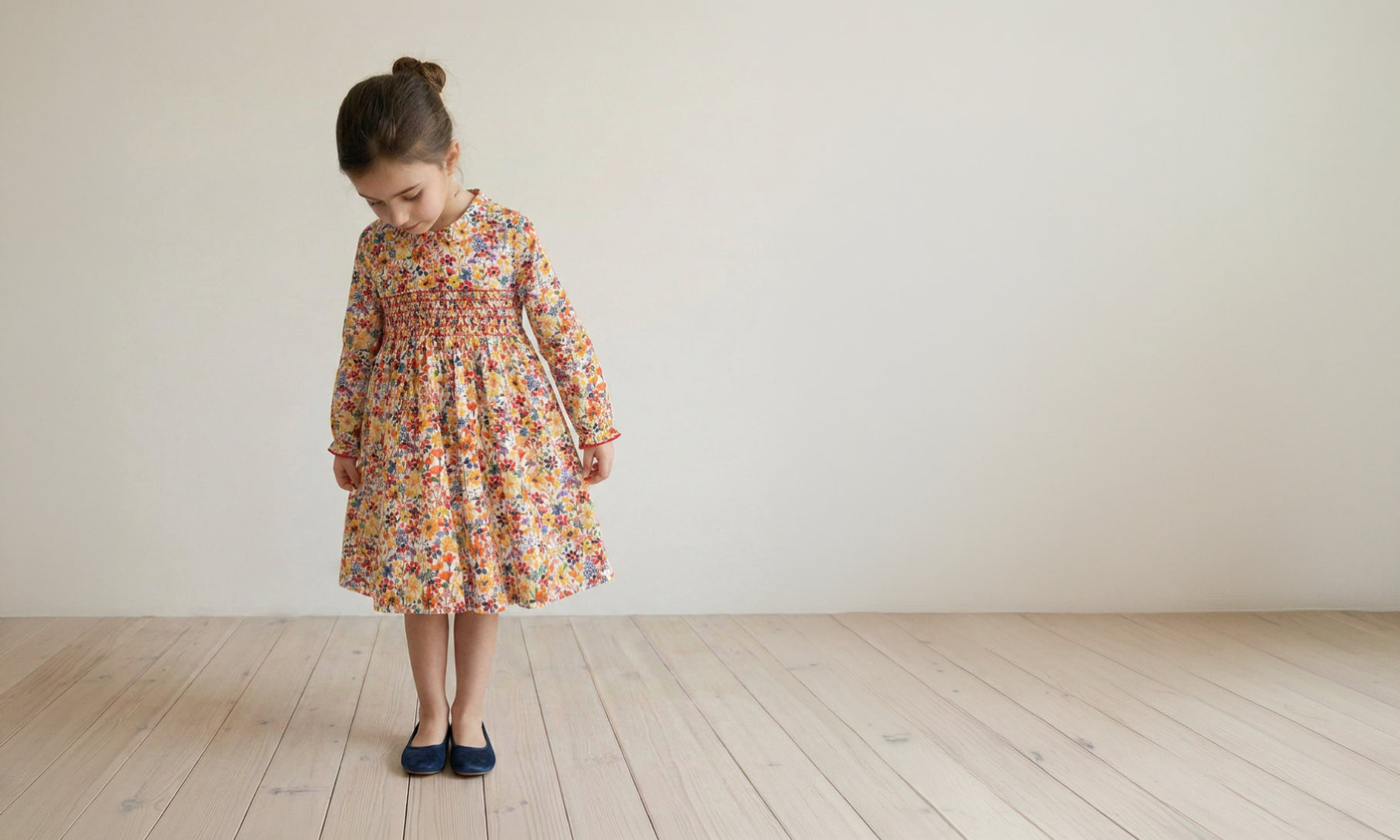 A young girl with her hair in a bun stands on a light wood floor against a plain off-white wall, wearing a long-sleeved floral smocked dress in orange, yellow, and blue tones, paired with dark blue shoes.