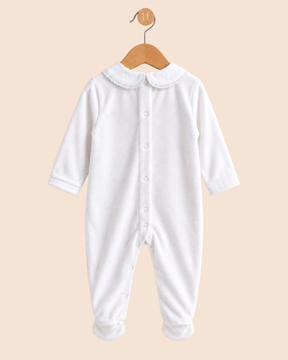 A white long-sleeve baby footed jumpsuit with a Peter Pan collar and snap-button front, hanging on a wooden hanger against a solid pale peach background.