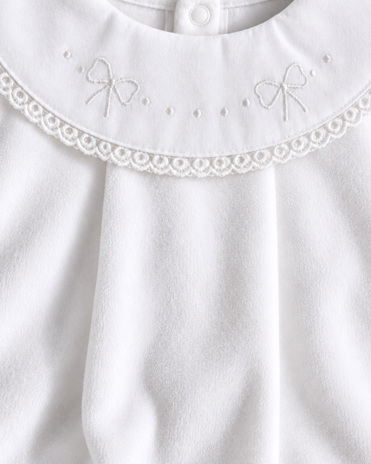 Close-up of a white baby garment featuring a decorative lace collar with embroidered bow details and a snap button at the neckline.