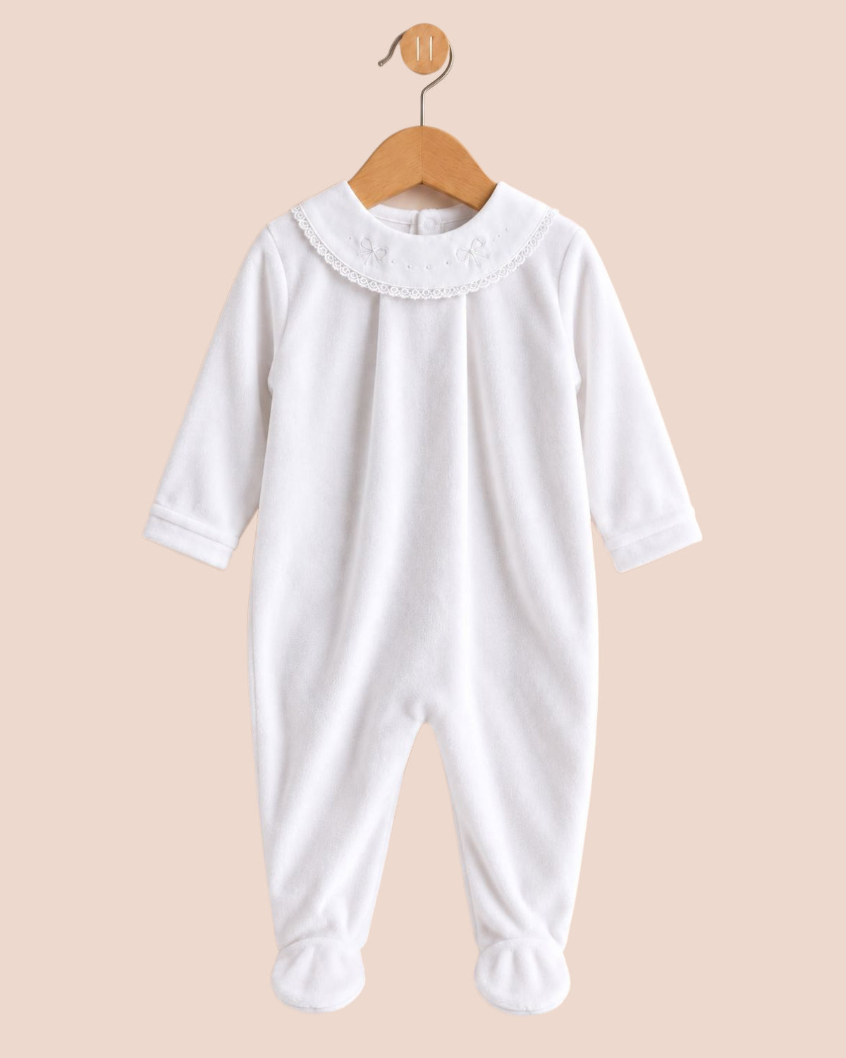 A white long-sleeve baby footed jumpsuit with a lace Peter Pan collar, hanging on a wooden hanger against a solid pale peach background.