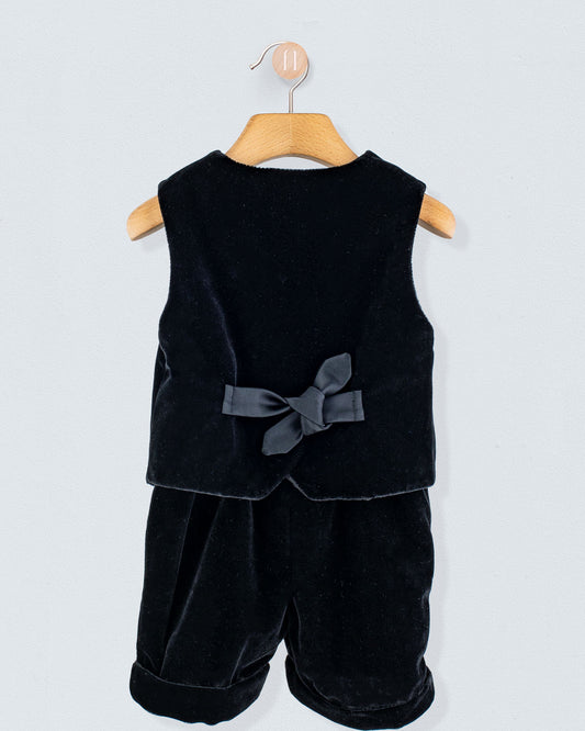 Adam 2-Piece Black Velvet Vest and Suspender Shorts on a hanger, showcasing the sleeveless vest with a back bow and matching shorts with cross-back suspenders.