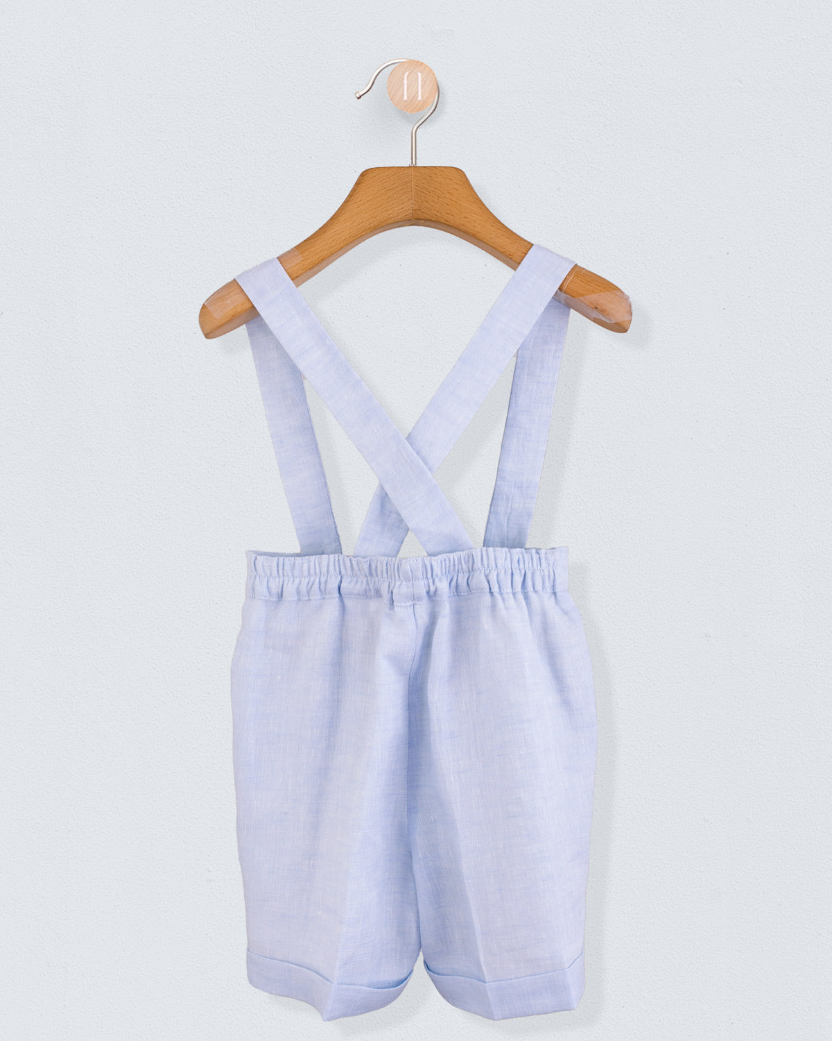 Back view of light blue linen suspender shorts with crossed straps and elastic waistband, hanging on a wooden hanger