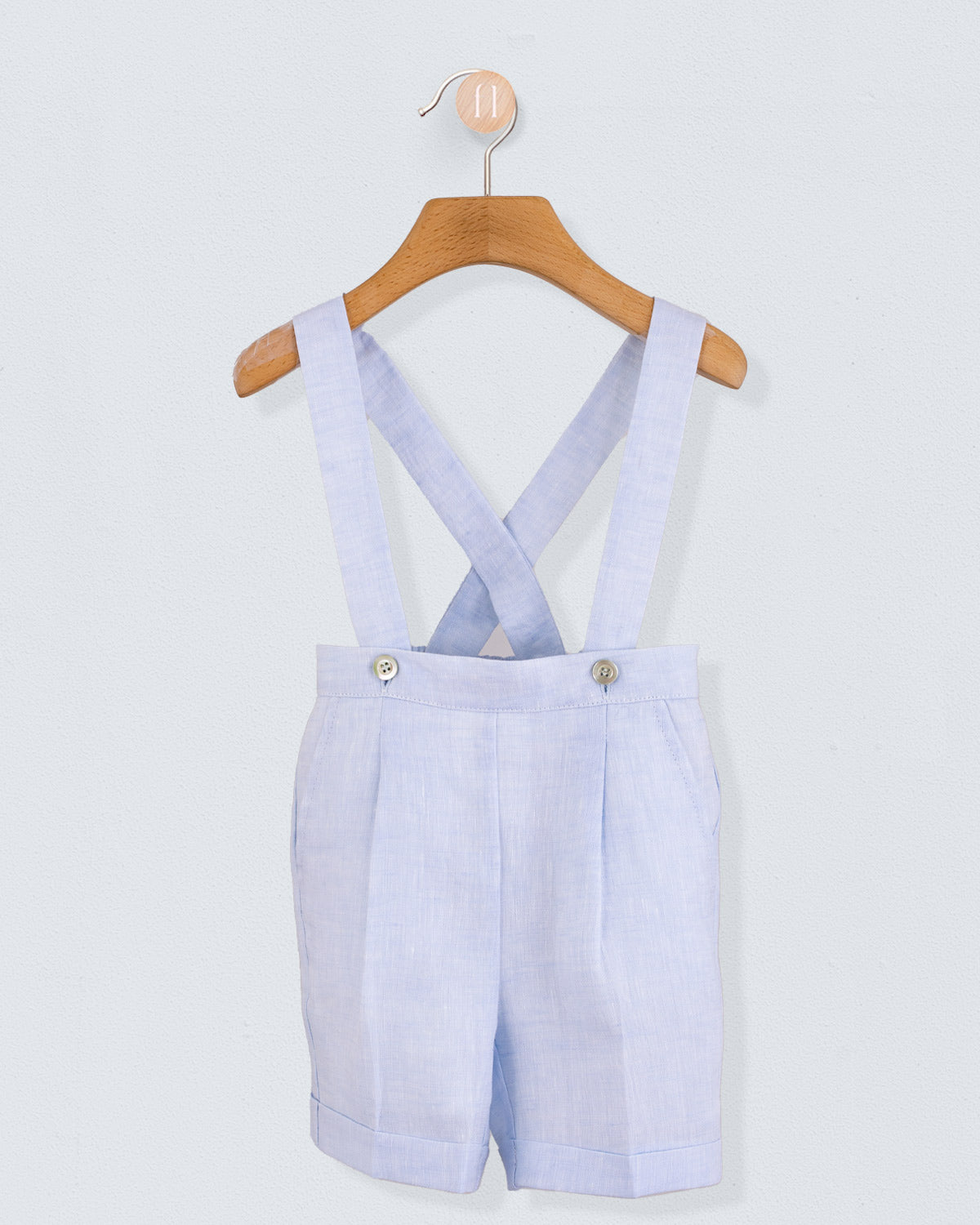 Front view of light blue linen suspender shorts with crossed straps buttoned at the waist, displayed on hanger.