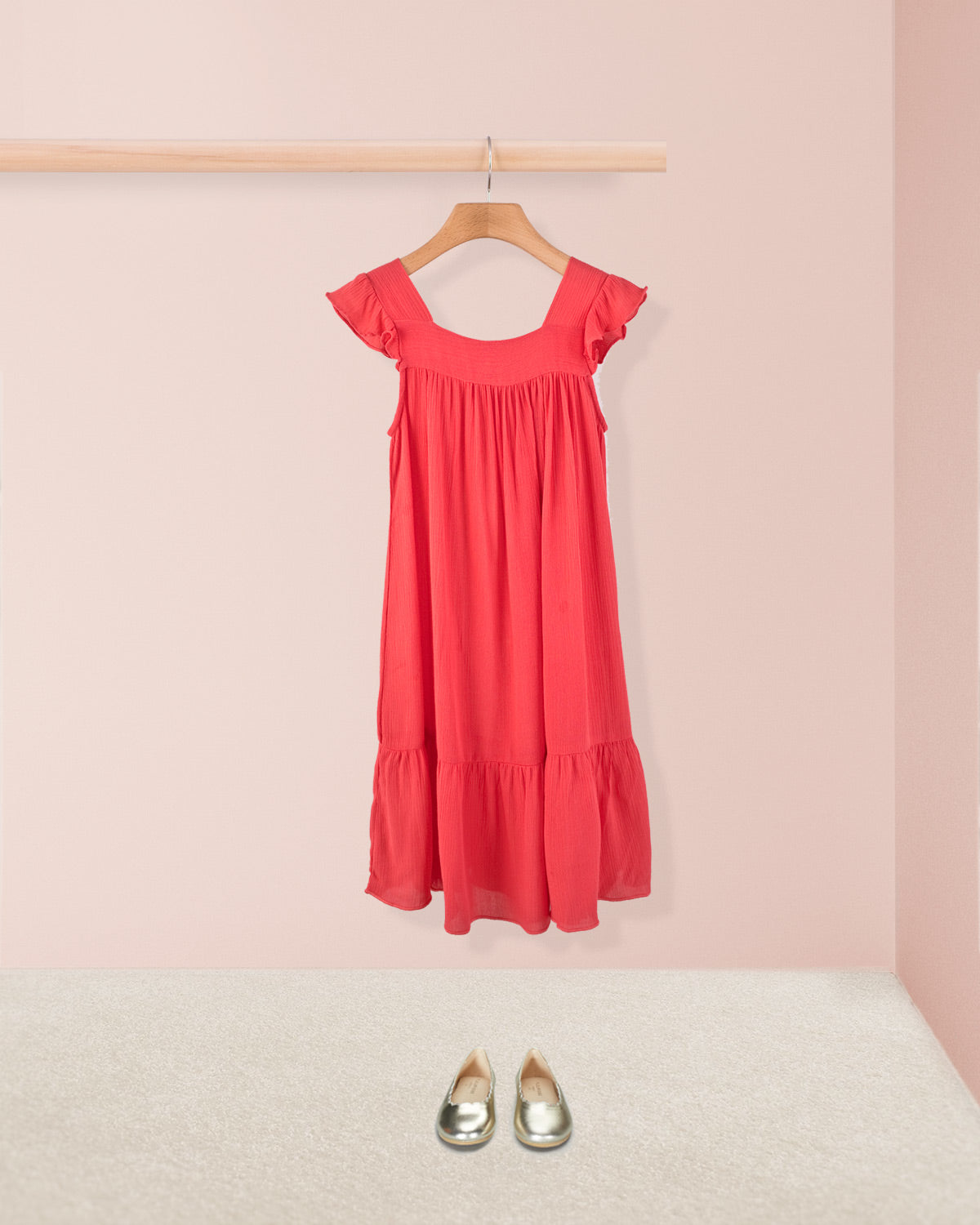Coral ruffle dress styled with metallic toddler shoes hanging on a wooden hanger. 