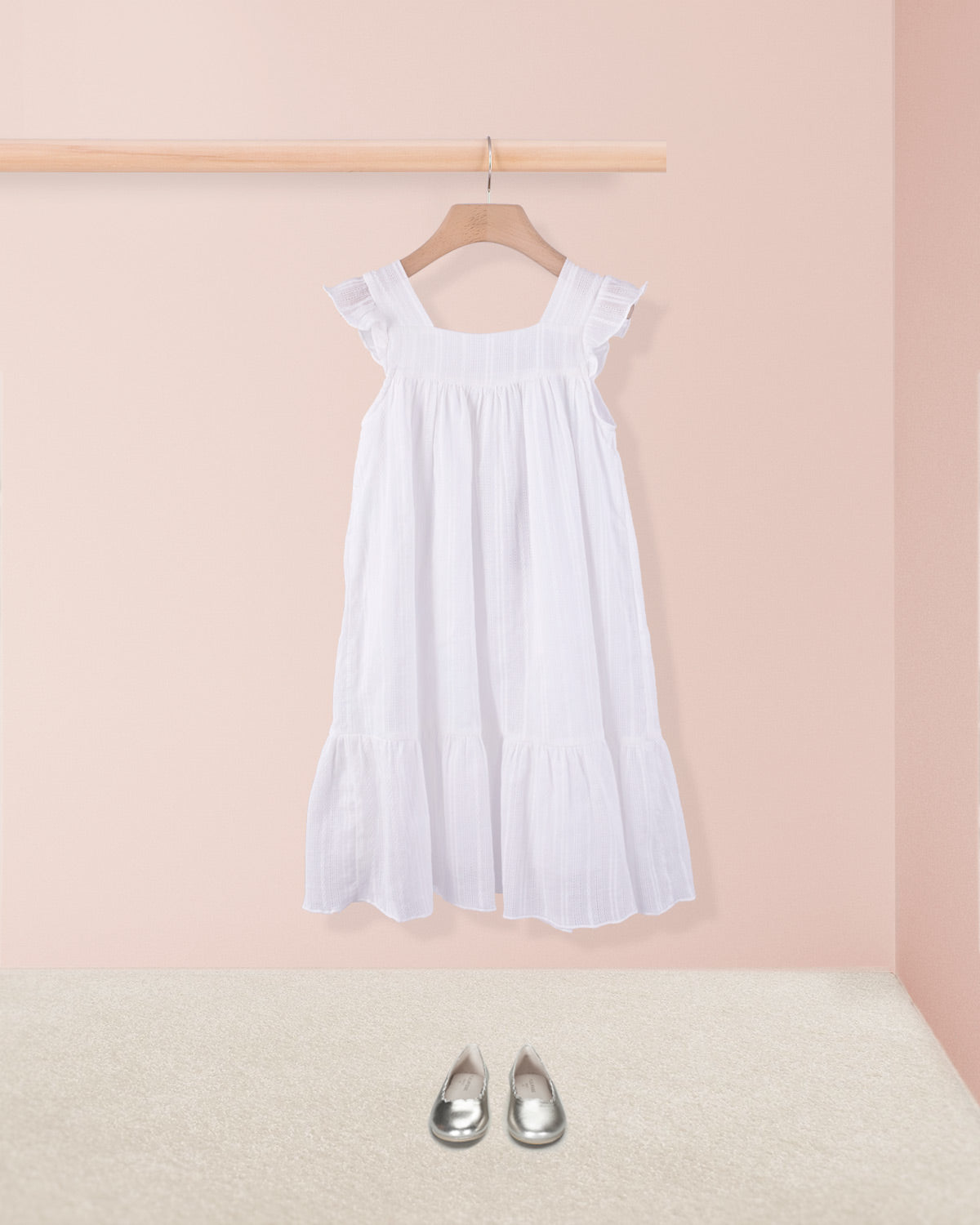 White ruffle dress styled with metallic toddler shoes hanging on a wooden hanger. 