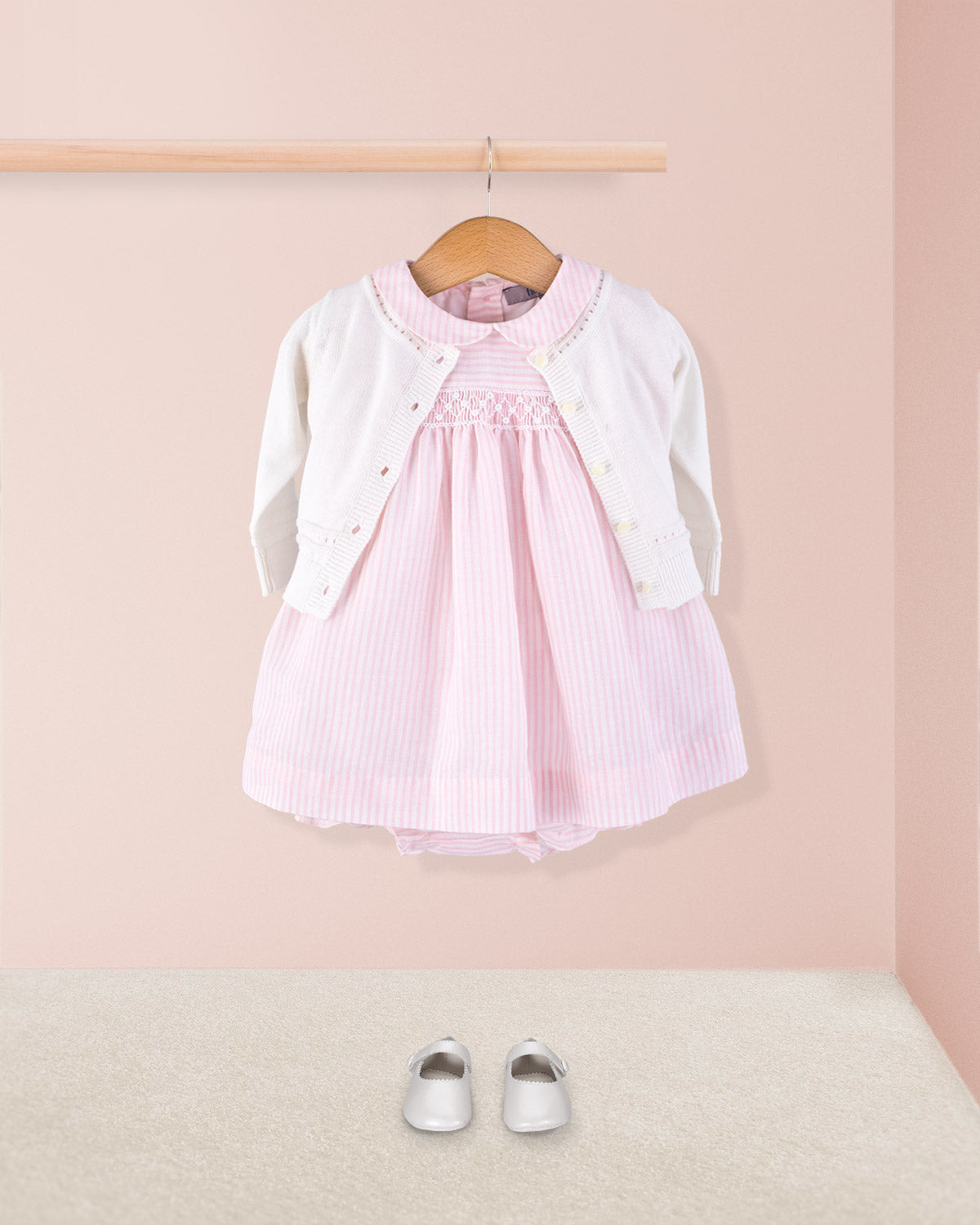 Baby Eva 2-piece pink striped dress paired with a white knit cardigan and white crib shoes, styled hanging on a wooden rod above a soft beige carpet.