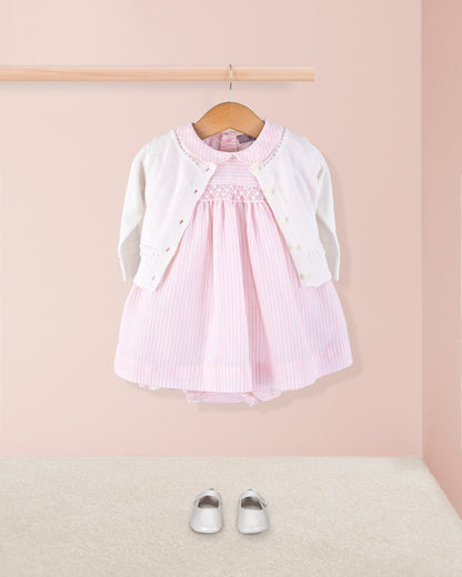 Baby Eva 2-piece pink striped dress paired with a white knit cardigan and white crib shoes, styled hanging on a wooden rod above a soft beige carpet.