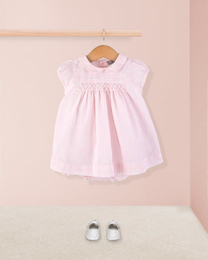 Front view of Baby Eva 2-piece dress paired with silver crib shoes, displayed hanging on a wooden rod above a carpeted floor for a full styled look.