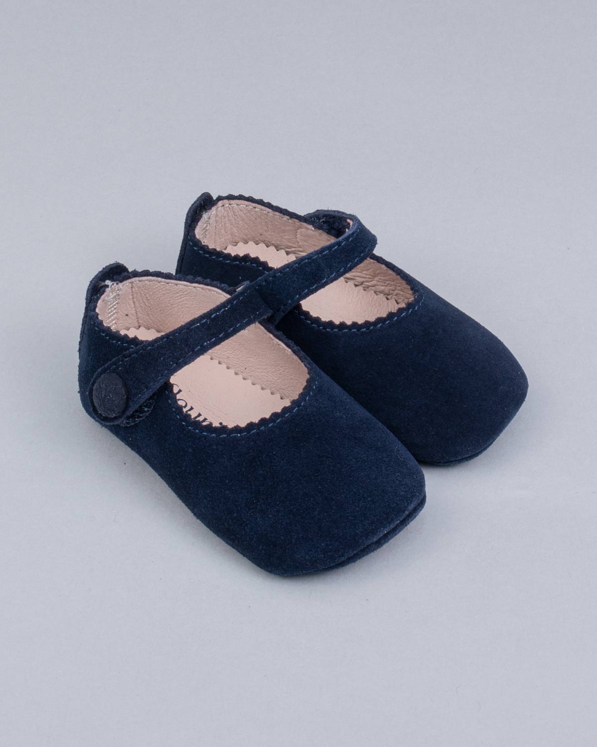 Pair of navy suede baby crib shoes with scalloped trim and velcro strap, featuring a decorative button, displayed on a soft grey background.