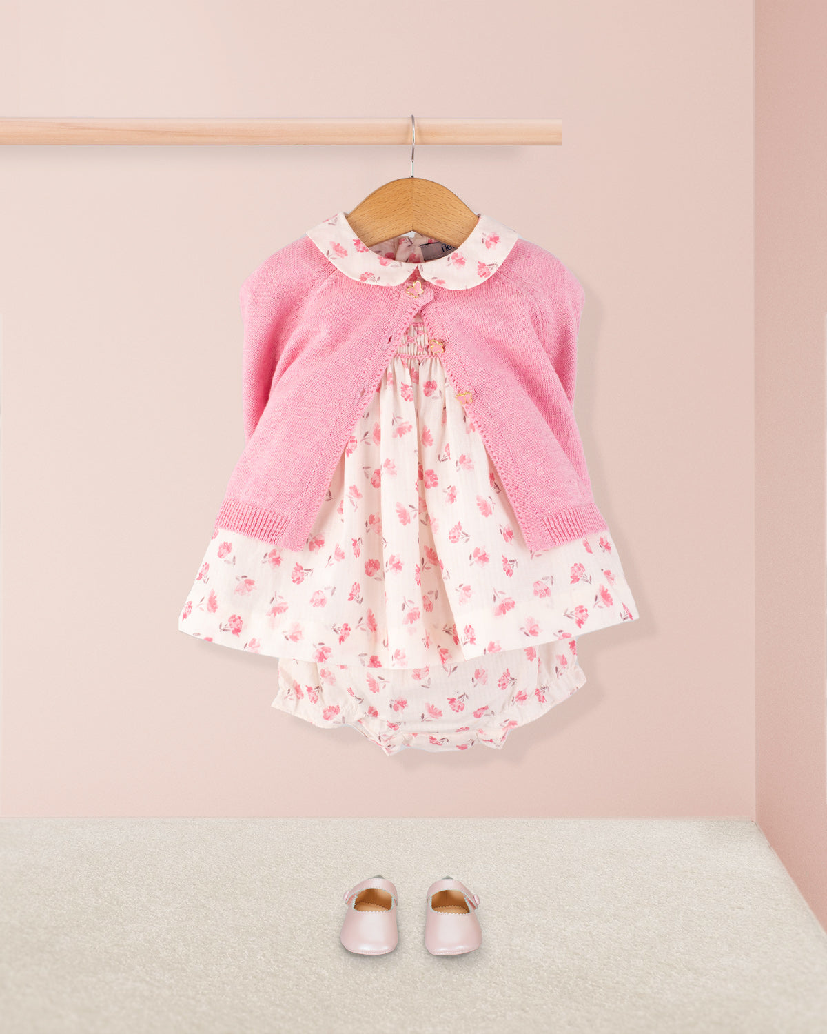 Baby Eva Versaille Rose Octuouse dress styled with a pink cardigan and matching shoes, displayed on a wall-mounted hanger above a soft carpeted floor.