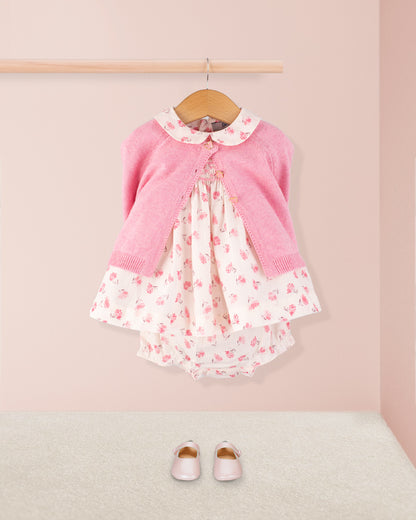 Baby Eva Versaille Rose Octuouse dress styled with a pink cardigan and matching shoes, displayed on a wall-mounted hanger above a soft carpeted floor.
