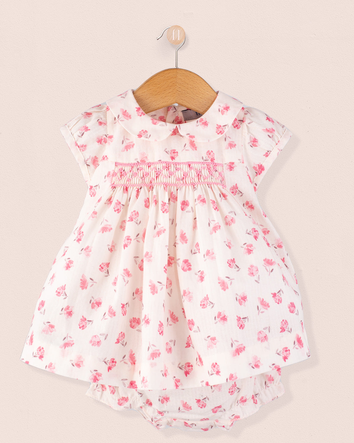Baby Eva Versaille Rose Octuouse dress set displayed on a hanger, showing the front of the white dress with pink floral print and smocking detail at the chest, matched with a diaper cover.