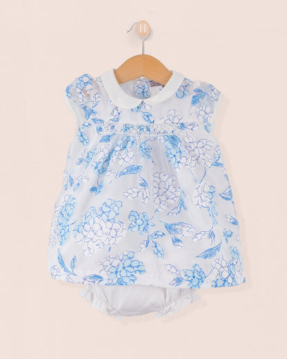 Front view of a sleeveless baby outfit in a blue and white floral print made of laser-cut cotton with a white Peter Pan collar. Matching bloomers visible underneath. Garment is hung on a wooden hanger against a pale pink wall.
