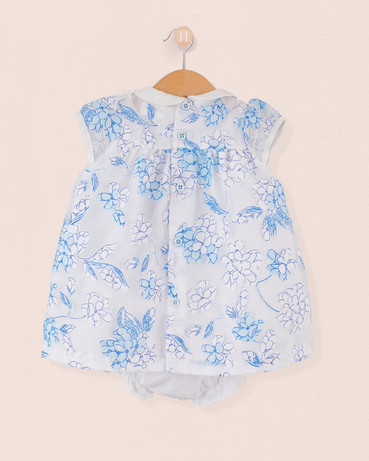 Back view of the same baby outfit in blue and white floral print with button closure along the yoke. Matching bloomers visible underneath. Garment is hung on a wooden hanger against a pale pink wall.