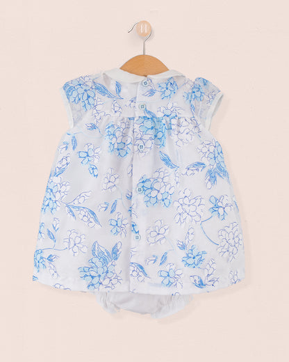 Back view of the same baby outfit in blue and white floral print with button closure along the yoke. Matching bloomers visible underneath. Garment is hung on a wooden hanger against a pale pink wall.