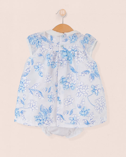 Back view of the same baby outfit in blue and white floral print with button closure along the yoke. Matching bloomers visible underneath. Garment is hung on a wooden hanger against a pale pink wall.